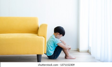 Unhappy Boy Feeling Guilty.Little Boy Feeling Guilty While Sitting And Hug The Knee And Bend Down The Head Behind The Sofa.Kid Feeling Guilty After Doing A Mistake.