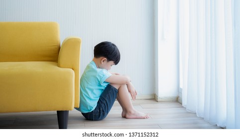 Unhappy Boy Feeling Guilty.Little Boy Feeling Guilty While Sitting And Hug The Knee And Bend Down The Head Behind The Sofa.Kid Feeling Guilty After Doing A Mistake.