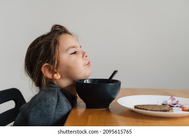 Unhappy Child Girl Eats Soup From Black Bowl With Bread And Onion. Lifestyle Photo Of Kid In Kitchen Having A Meal, Screaming Kid. Picky Eater