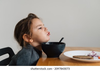 Unhappy Child Girl Eats Soup From Black Bowl With Bread And Onion. Lifestyle Photo Of Kid In Kitchen Having A Meal, Screaming Kid. Picky Eater