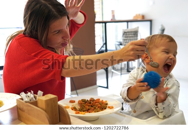 母親がいたずらで幼児をだますようにしている間 白い背景を持つレストランでの食事を拒否した不幸な子ども とにかく彼は食べたくない の写真素材 今すぐ編集