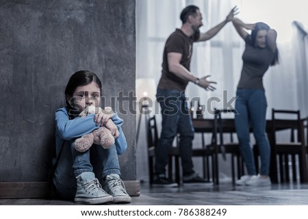 Unhappy childhood. Nice unhappy cheerless girl sitting on the floor and holding her toy while listening to her parents fight