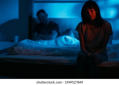 Unhappy Couple. Domestic Violence. Female Abuse. Scared Insecure Humiliated Woman Sitting On Bed On Distance From Aggressive Man In Dark Night Red Blue Neon Light On Defocused Background.