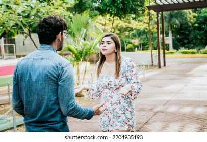 Unhappy Couple Standing Arguing In A Park. Disgusted Teenage Couple Arguing In A Park, Man And Woman Arguing In A Park. Young Couple Arguing Misunderstanding In A Park