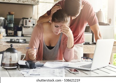 Unhappy Couple Unable To Pay Loan On Time: Stressed Female Doing Paperwork Sitting At Table With Laptop, Papers, Calculator And Cell Phone. Man Trying To Support His Wife, Patting Her On Head