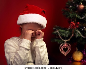 Unhappy Crying Kid Girl In Santa Claus Hat Near The Christmas Tree Because Did Not Get A Gift. Closeup Portrait On Red Background