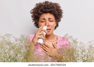 Unhappy Curly Haired Woman Suffers From Runny Nose Uses Nasal Spray Has Red Swelling Eyes Has Allergic Rhinitis Reaction On Wild Plant Poses Against White Background. People And Sickness Concept