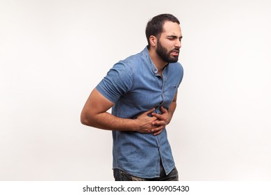 Unhappy Depressed Man With Beard Holding Hands On Stomach Feeling Acute Pain, Suffering Indigestion And Nausea, Duodenal Ulcer. Indoor Studio Shot Isolated On White Background