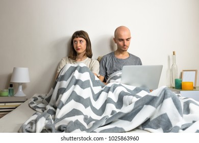 Unhappy or dissatisfied young couple in bed. Man and woman sit in modern bedroom after quarrel or dispute, girl depressed or sad, male with laptop computer