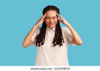 Unhappy Distressed Woman With Black Dreadlocks Has High Blood Pressure, Rubs Temples To Relieve Headache, Keeps Eyes Shut, Has Unbearable Migraine. Indoor Studio Shot Isolated On Blue Background.