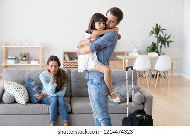 Unhappy Diverse Family In Living Room At Home. Sad Father Hugs Hold On Hand Small Daughter, Mother Sitting On Sofa. Parents Divorcing, Child Stay With Mommy Dad Leave With Suitcase. Break Up Concept