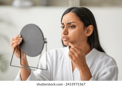 Unhappy eastern young woman in white robe looking at mirror, checking skin, touching her chin, home interior. Acne, pimples, dull skin concept, closeup
