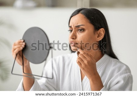 Unhappy eastern young woman in white robe looking at mirror, checking skin, touching her chin, home interior. Acne, pimples, dull skin concept, closeup