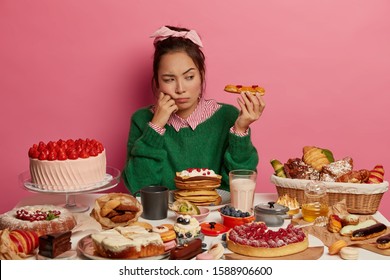 Unhappy Ethnic Girl Looks With Temptation At Delicious Dessert, Sad As Can Put On Weight After Eating High Caloried Food, Has Variety Confectionery And Pasrty On Table, Poses In Room With Pink Walls