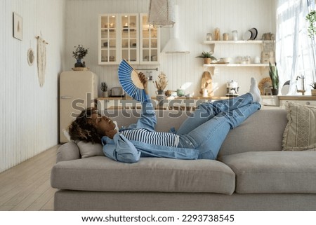 Unhappy exhausted and overheated young African woman lying on sofa at home waving fan to cool down, black female having hot flashes, feeling hot during heatwave in apartment without air conditioning