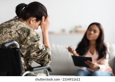 Unhappy Female Soldier With Disability Attending Therapy Session With Black Psychiatrist Lady, Military Woman In Camouflage Uniform Sitting In Wheelchair And Touching Head, Suffering Depression