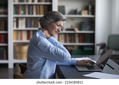 Unhappy frustrated senior woman feeling strong neck ache after using laptop computer at uncomfortable workplace in library, suffering from inflammation, muscles tension. Elderly healthcare concept