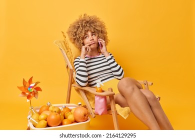 Unhappy Frustrated Woman Has Phone Conversation With Friend Looks Very Sad Poses In Comfortable Deck Chair Spends Summer Vacation At Beach Isolated Over Vivid Yellow Background. Holidays Concept