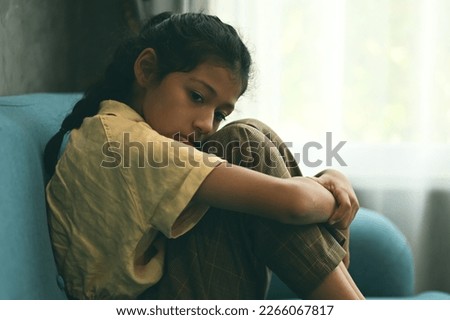 Unhappy girl portrait, Sad child sitting on sofa at home, Upset girl hugging knees alone, Concept of lonely girl and kid with trouble and violence