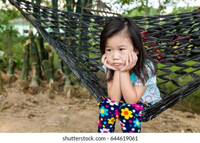 Unhappy Girl Propping Up Her Face And Sitting Alone In Hammock. Asian Child Bored, Frustrated And Fed Up, Kid Emotion. Concept For Depression Stress Or Frustration. Outdoors On Summer Day.