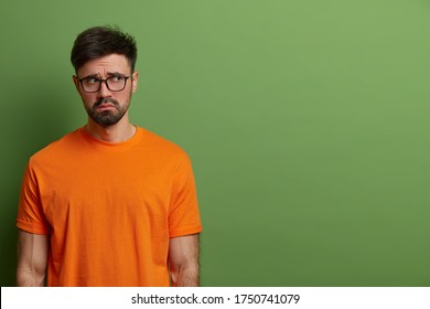 Unhappy gloomy young European man looks upset and disappointed, wears casual orange t shirt and spectacles, feels uneasy and moody, stands against green background, copy space for your promotion. - Shutterstock ID 1750741079