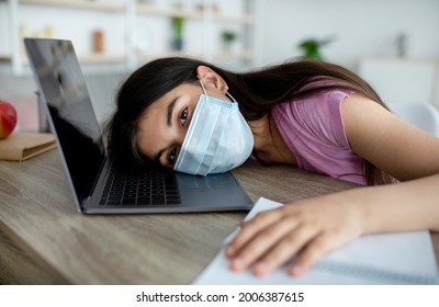 Unhappy Indian Teen Girl In Face Mask Lying On Laptop Keyboard, Feeling Bored Of Online Studies During Covid Quarantine. Hispanic Teenager Sick And Tired Of Distance Education, Indoors