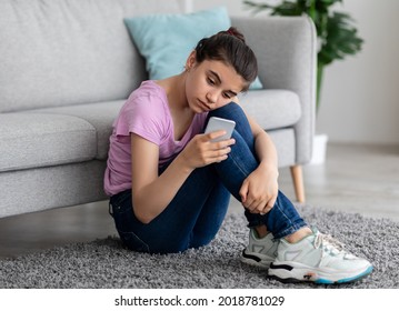 Unhappy Indian Teen Girl With Mobile Phone Feeling Lonely And Sad, Suffering From Cyber Bullying Online, Sitting On Floor In Living Room. Stressed Teenager Being Victim Of Internet Abuse