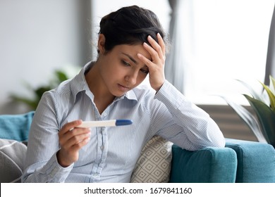 Unhappy Indian woman looking at pregnancy test close up, upset by result, stressed thoughtful young female touching forehead, sitting on couch, unwanted pregnancy or health problem, infertility