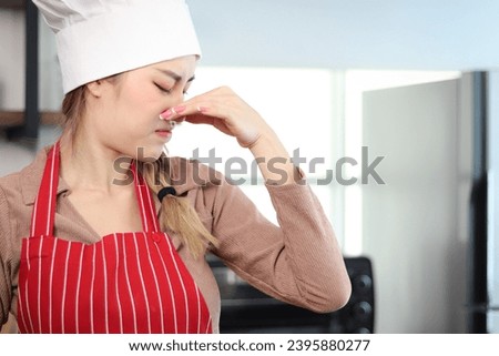 Unhappy irritated glamour Asian beautiful woman with apron chef and hat putting fingers to cover her nose at kitchen. Young female chef cannot tolerate bad smell and pungent while cooking food.
