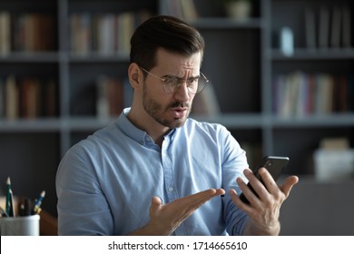 Unhappy irritated young man wearing glasses having problem with broken or discharged smartphone, indignant angry confused businessman looking at phone screen, reading bad news in message - Shutterstock ID 1714665610