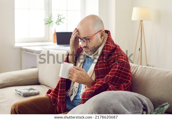 Unhappy joyless sick man at home treats flu and colds\
with medicinal drink with unpleasant taste. Middle-aged man wrapped\
in warm scarf and plaid looks at cup in his hands, not wanting to\
drink it.
