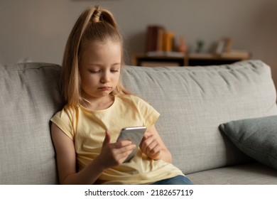 Unhappy Little Girl Using Mobile Phone Networking In Social Media Sitting On Couch At Home. Bored Kid Playing Game On Cellphone. Mobile Application, Gadgets And Kids Concept