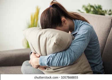Unhappy lonely depressed woman at home, she is sitting on the couch and hiding her face on a pillow, depression concept