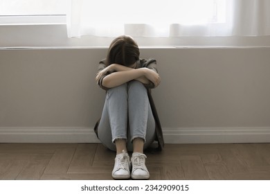 Unhappy lonely stressed young teen girl sitting on floor at wall, crying, bending head, covering face with arms, keeping close posture, coping with depression, fatigue, frustration, loss