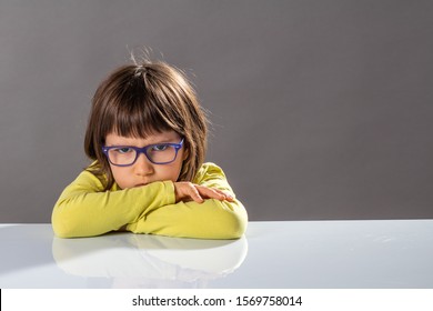 Unhappy Mad Child With Dirty Look Behind Eyeglasses And Arms Folded On Table Expressing An Irritated Pouting No