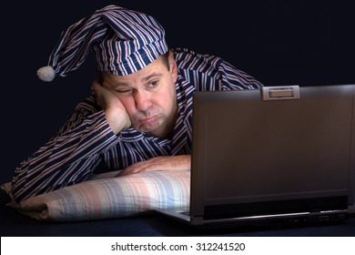 Unhappy Man With Computer In Bed. Sleepy Man In Striped Pajamas Lying In Bed With A Computer. A Man With Night Cap Lying With A Laptop. Bored Man In Pajamas In Bed With Computer.