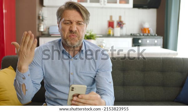Unhappy man feels uncomfortable while using\
smartphone. Angry man having problem with cell phone texting. The\
male user sitting on the sofa at home was frustrated by what the\
smartphone saw.