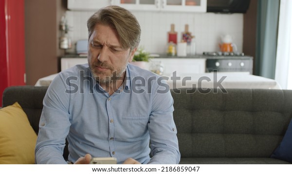 Unhappy man feels uncomfortable while using\
smartphone. Angry man having problems with cell phone messages. The\
male user sitting on the sofa at home gets very angry with what the\
smartphone sees.