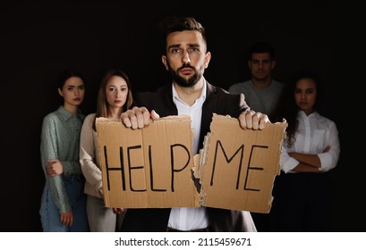 Unhappy Man With HELP ME Sign And Group Of People Behind His Back On Dark Background