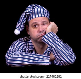 Unhappy Man In Pajamas On A Black Background. Frustrated Sleeper With A Night Cap Suffers From Insomnia. 