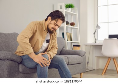 Unhappy man sitting on sofa at home and touching hurt knee with grimace of pain and suffering. Young guy with rheumatism, arthritis or osteoarthritis feeling intense severe pain in knee joint
