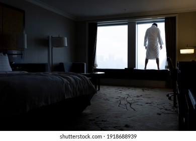 Unhappy man standing in window of hotel room, looking down to street and thinking about committing suicide. Male figure in bathrobe in backlight. Despair and depression concept.