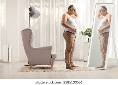 Unhappy mature man holding his big belly and looking at a mirror at home
