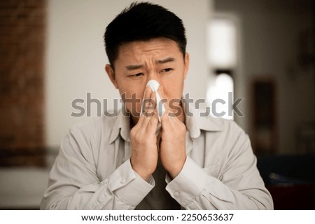 Unhappy middle aged asian man suffering from fever and flu, blowing nose in napkin, sitting in living room interior. Covid-19 lockdown, treatment of illness, cold and runny