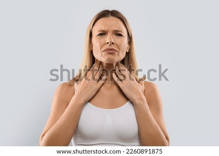 Unhappy middle aged blonde woman touching her neck, suffering from pain in throat, checking enlarged adenoids isolated on grey studio background, copy space. Sore throat concept