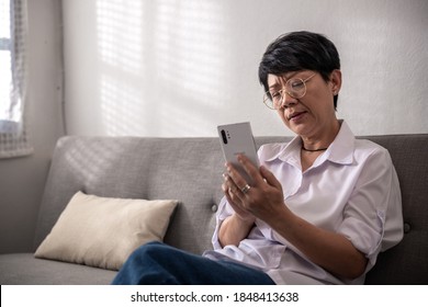 Unhappy Middle-aged gray-haired Asian woman has questions while sitting alone texting on the mobile phone or shopping online. A retired woman on the sofa at home. And senior retired Concept.