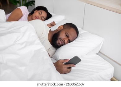 Unhappy millennial african american husband looks at phone, wife sleeps next to him on bed in bedroom interior. Relationship problems, gadget addiction, online game, bad news, insomnia and treason