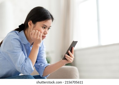 Unhappy Millennial Asian Woman Sitting On Couch In White Living Room, Using Smartphone At Home, Leaning On Her Arm, Looking At Cell Phone Screen, Scrolling On Social Media, Feeling Bored, Copy Space