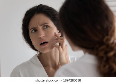 Unhappy millennial Caucasian woman look in mirror in bathroom distressed with dry scaling skin. Upset confused young female frustrated by facial pimple or first wrinkles. Beauty procedure concept. - Shutterstock ID 1894280638