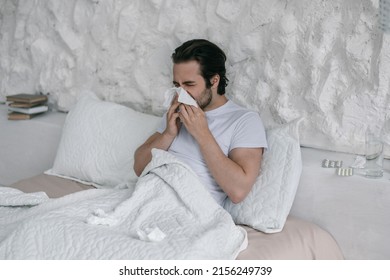 Unhappy Millennial European Male Lies On Bed, Blows Nose At Napkin In White Bedroom Interior. Runny Nose, Disease Treatment Alone, Flu And Cold, Health Problems At Home During Covid-19 Quarantine